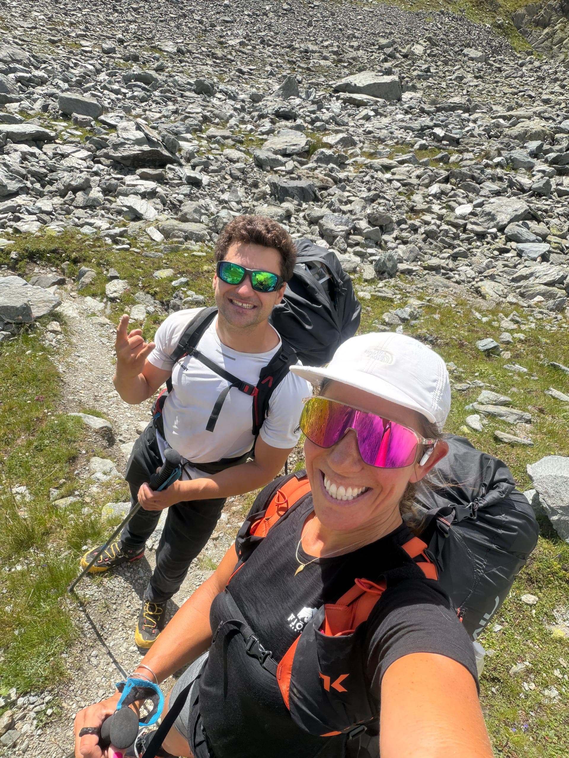 Un alpiniste et une alpiniste en selfie sur un sentier de moraine, parapentes sur le dos, avant le Grand Combin