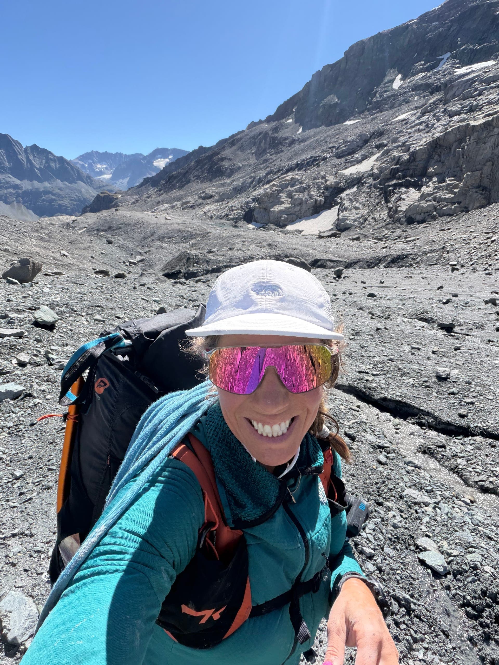 Une alpiniste souriante en selfie sur un terrain de pierres, avec une grande paroi rocheuse derrière