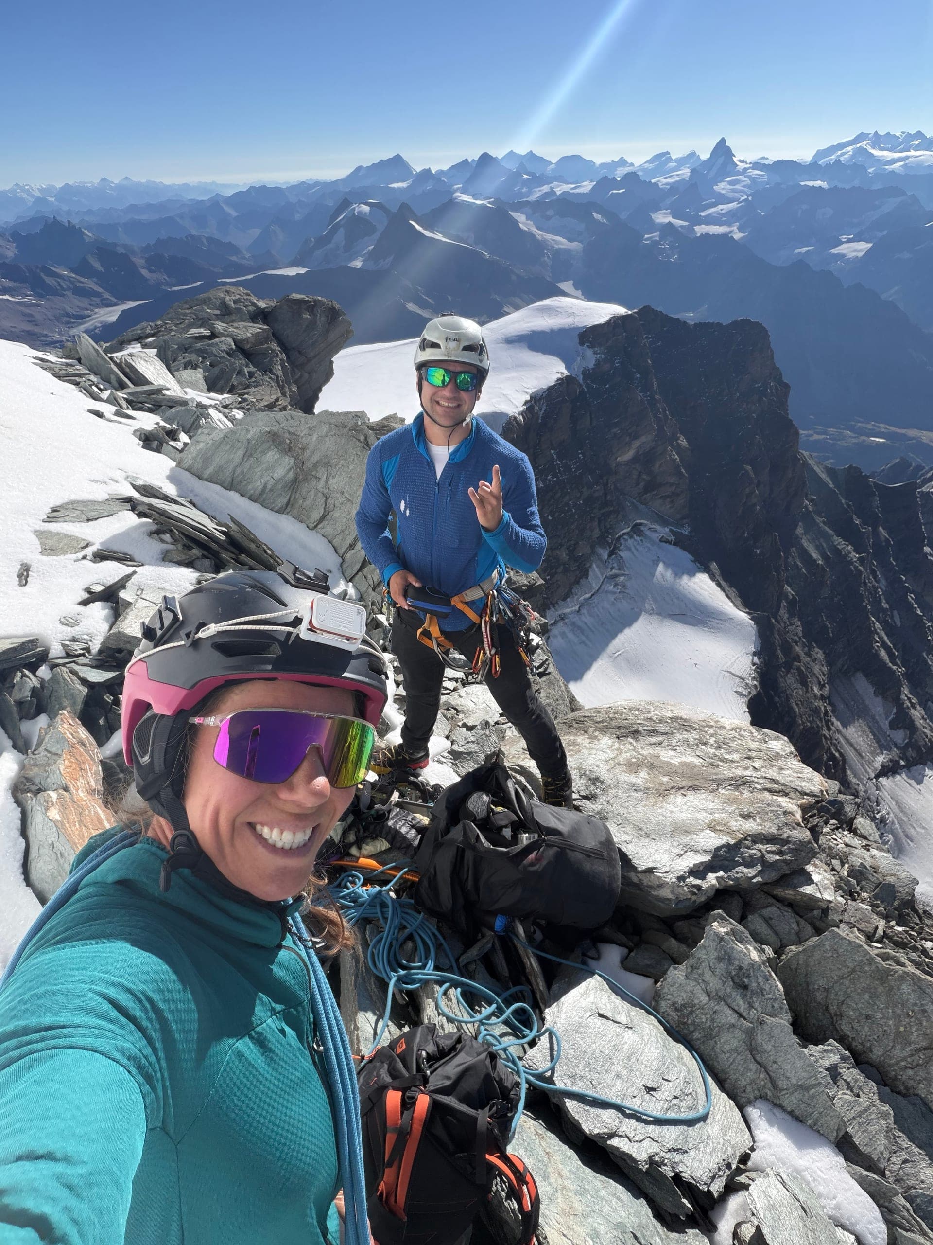 Une alpiniste en selfie au sommet, un compagnon de cordée derrière elle, arêtes et glaciers en contrebas