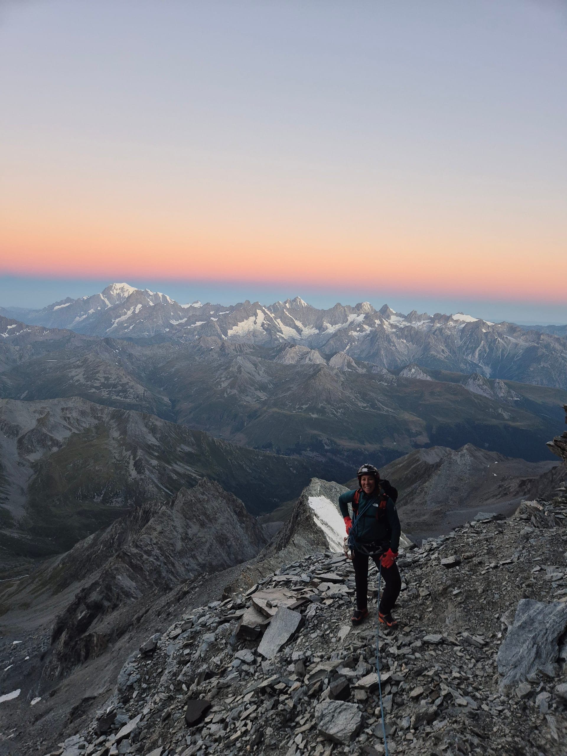 Un alpiniste sur une arête de pierres au lever du jour, ciel orangé et chaîne de montagnes au loin