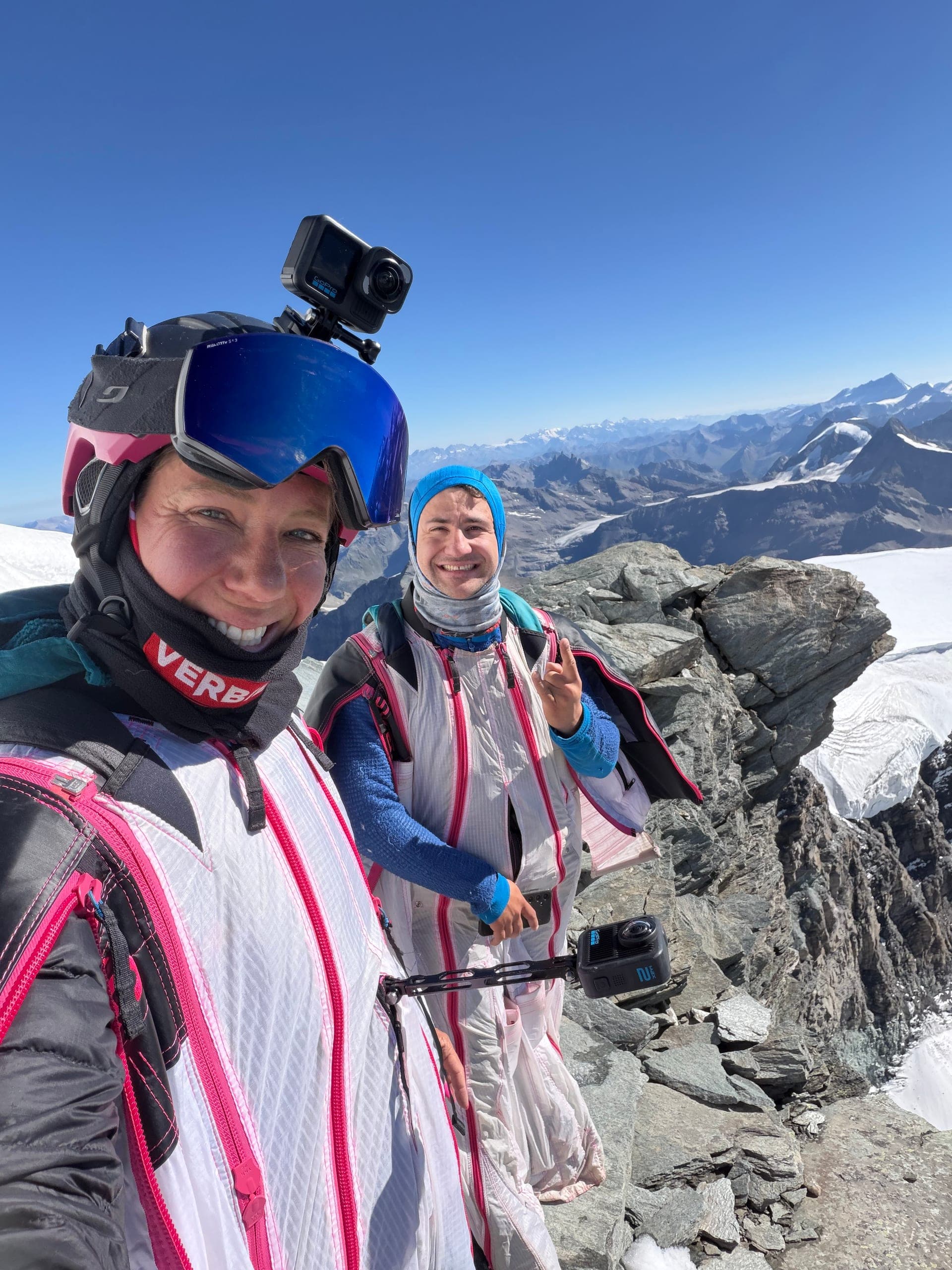 Deux wingsuiters en combinaisons colorées, caméra sur le casque, en selfie sur un sommet rocheux avant le vol