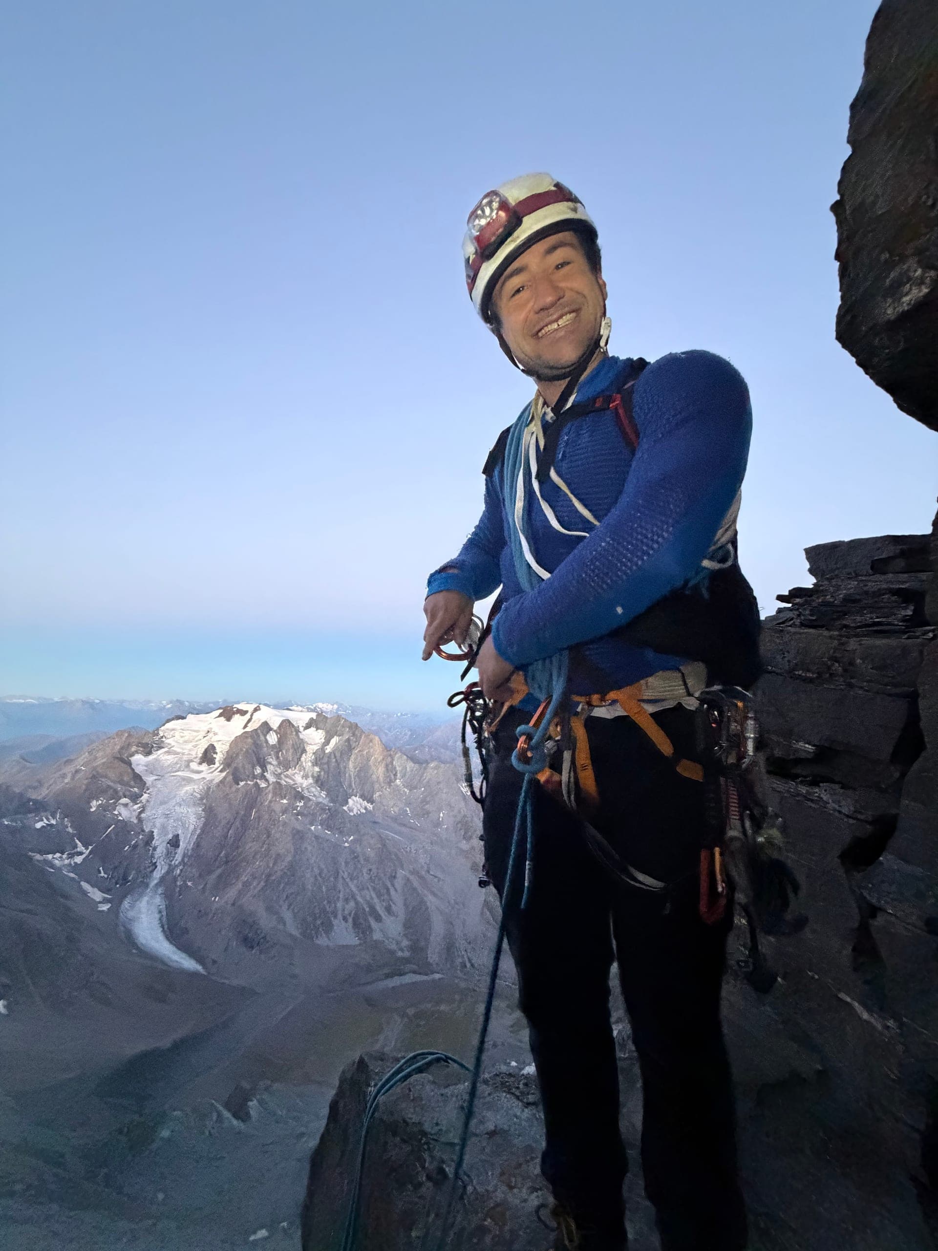 Un alpiniste encordé, casque sur la tête, à l’aube, un sommet englacé derrière lui