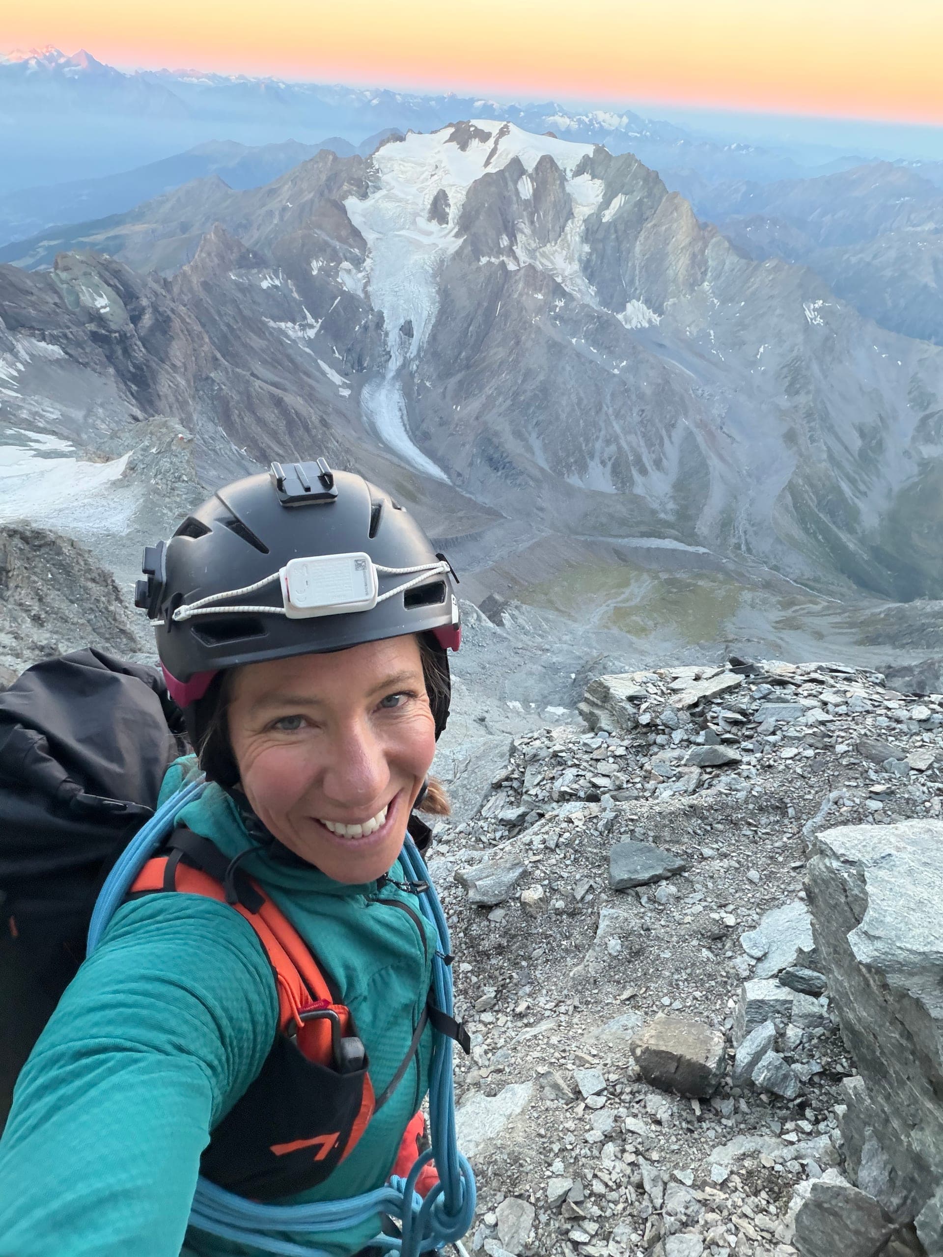 Une alpiniste souriante, lampe frontale sur le casque, au lever du jour, un massif englacé derrière elle