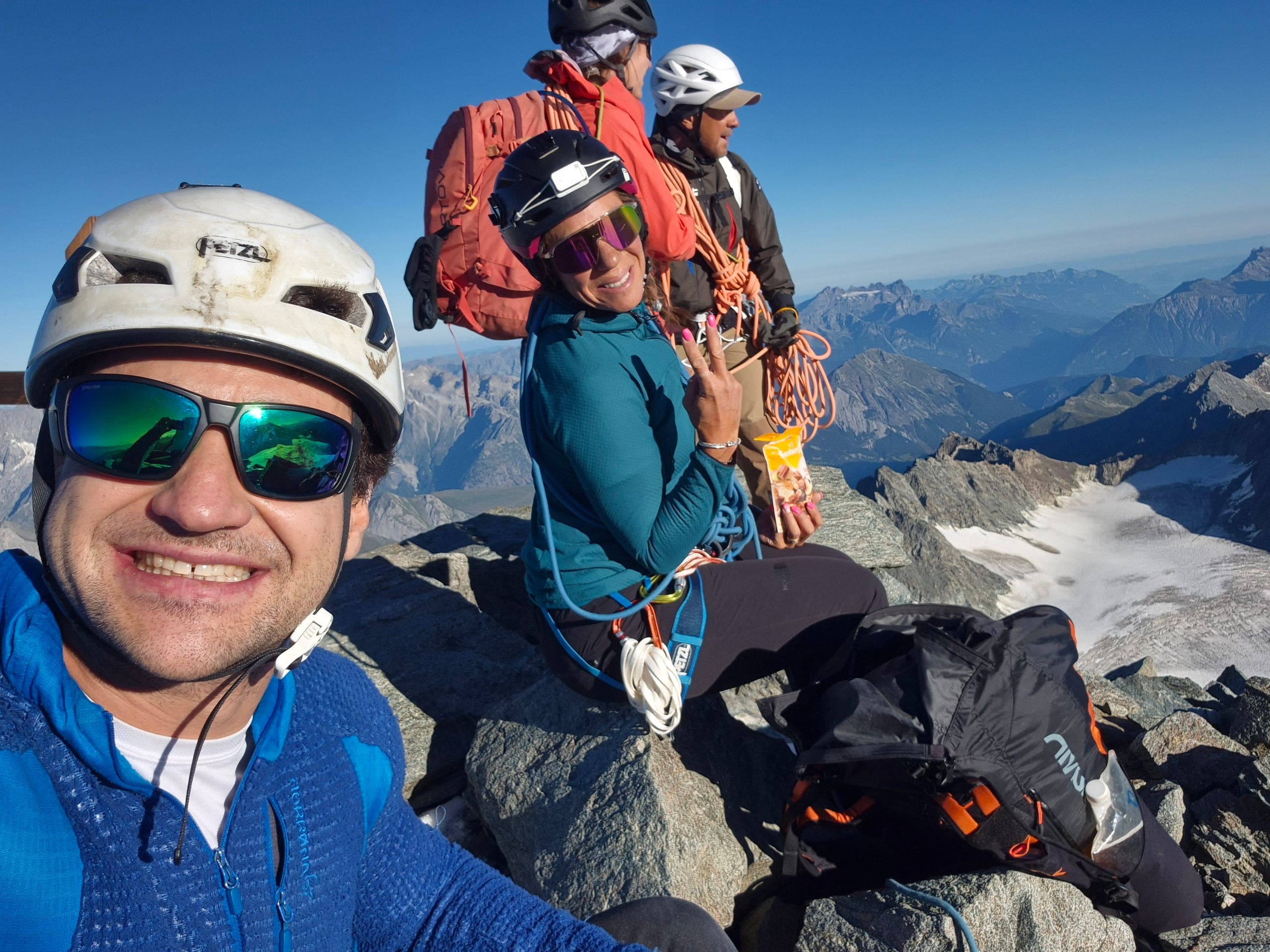 Un alpiniste en selfie, une alpiniste et un autre grimpeur assis derrière lui avec une voile de wingsuit, au sommet