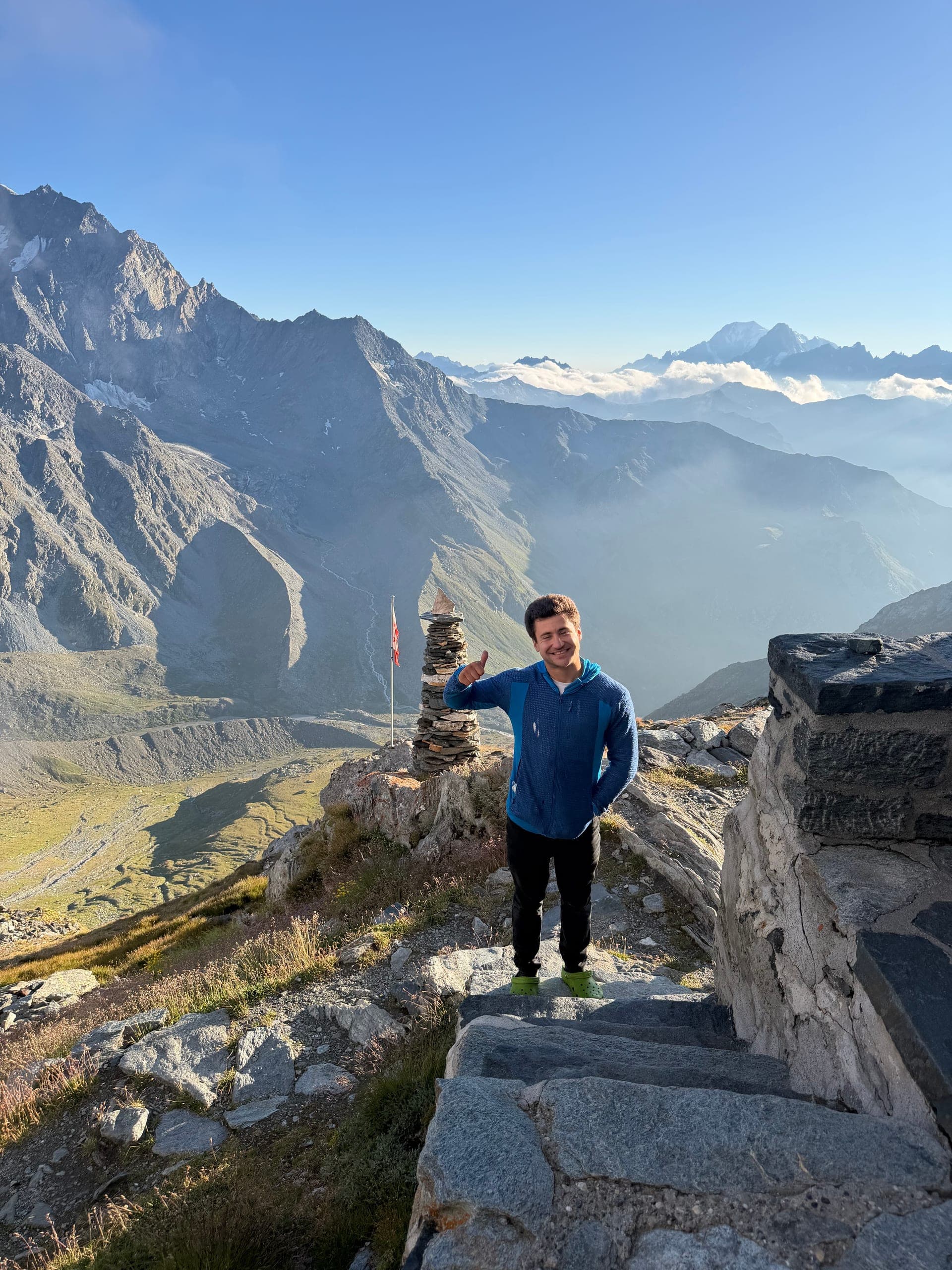 Un alpiniste en bleu debout sur des marches de pierre près d’un cairn, vallée et montagnes derrière