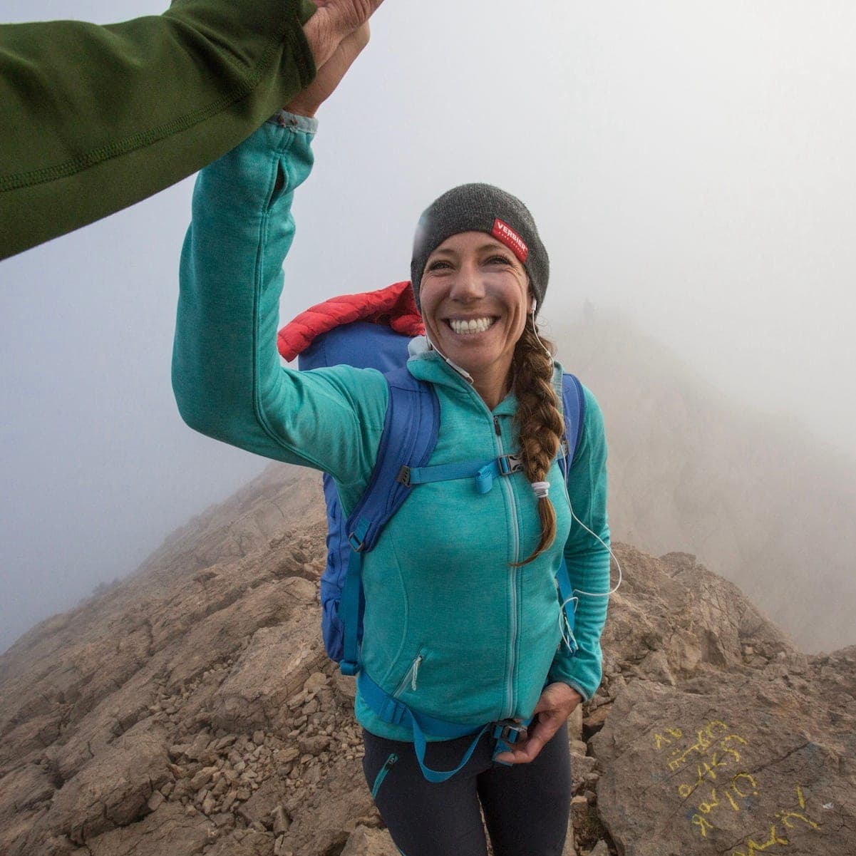 A smiling climber in a teal fleece with a backpack, in the mist on the Bisotoun ridge