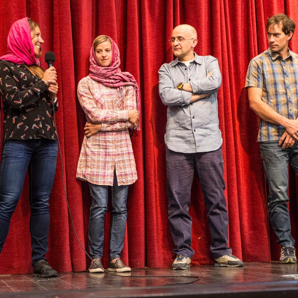 Four people standing before a red curtain on a stage, one speaking into a microphone, in Iran