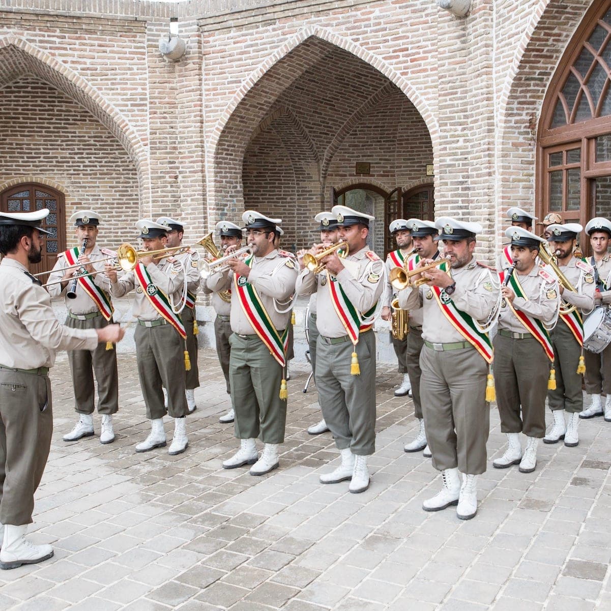 A brass band in beige uniforms and tricolor sashes playing in a brick arcaded courtyard, Iran