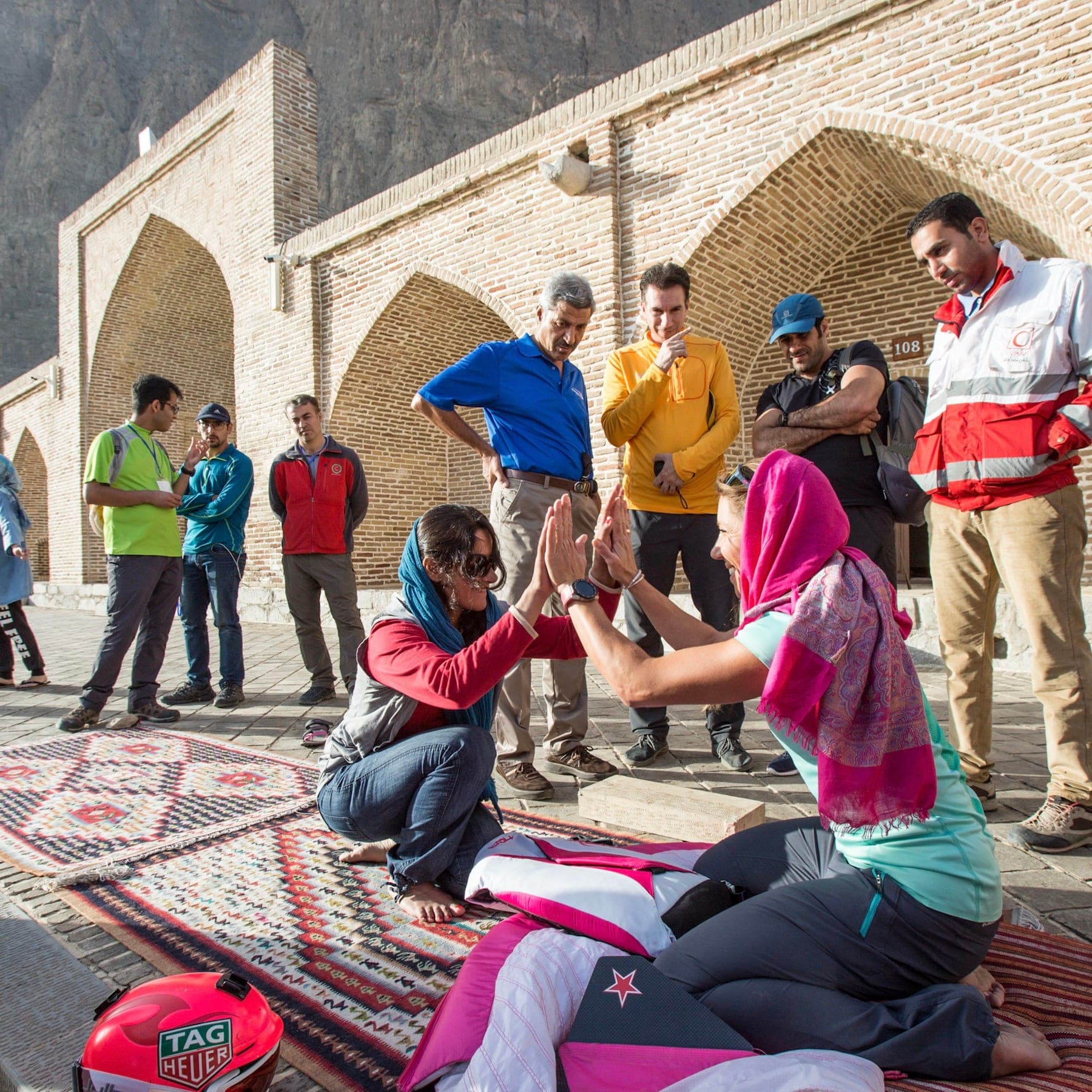 Two women in headscarves working on a folded wing on a rug while a group gathers under the arcades, Iran