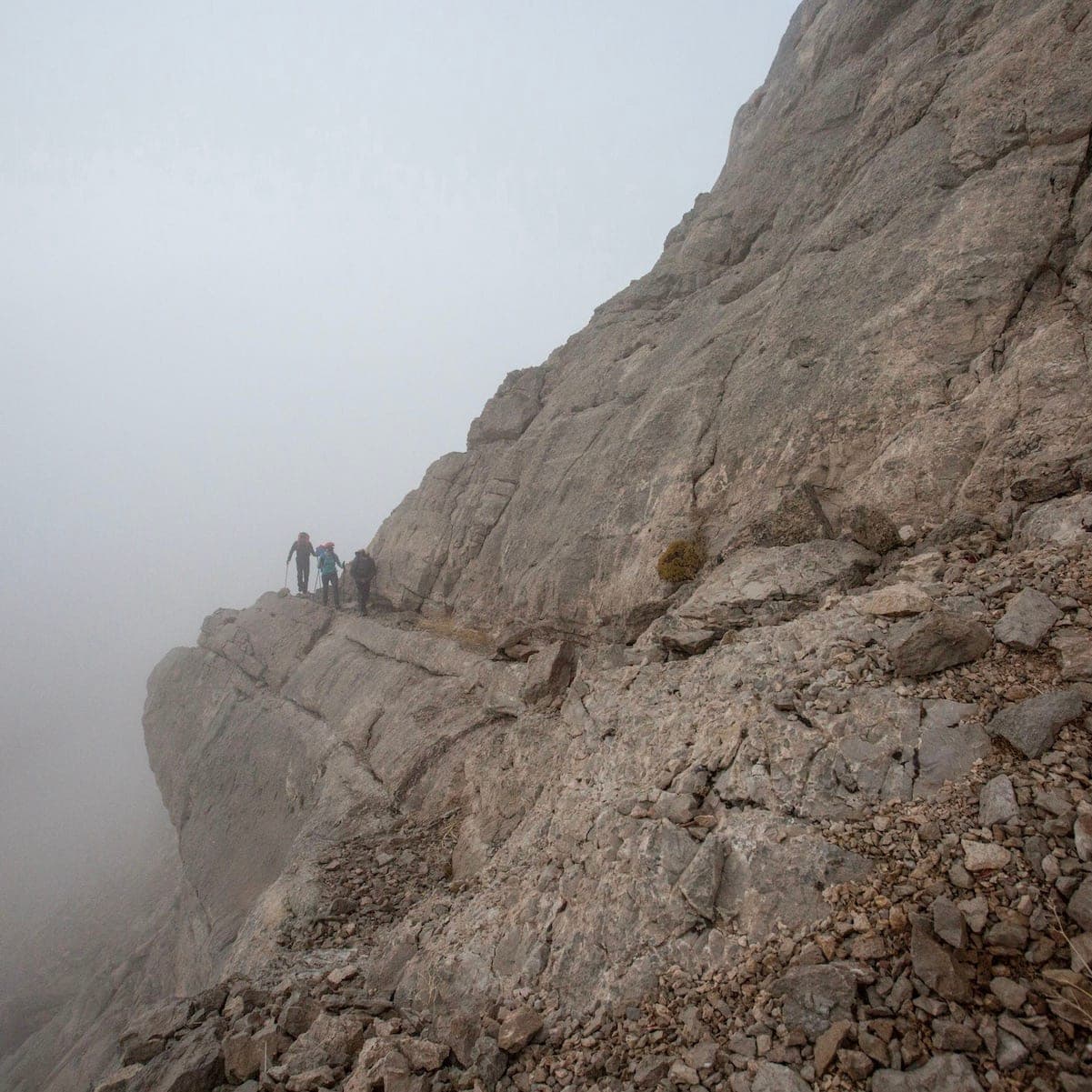 Rock face lost in mist, with a few tiny climbers on a ledge path, Bisotoun