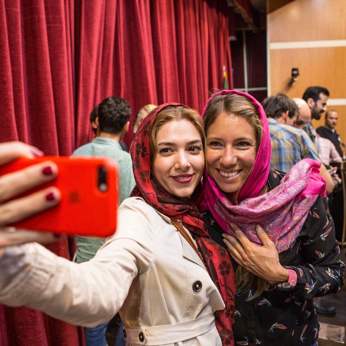Two women in headscarves taking a selfie in front of a red curtain, in Iran