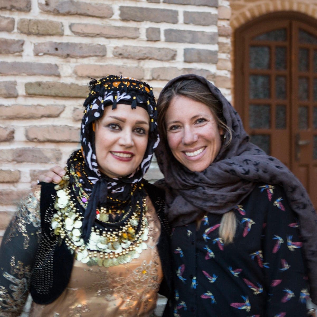 Two women smiling arm in arm in front of a brick wall, one in embroidered traditional dress with gold coins