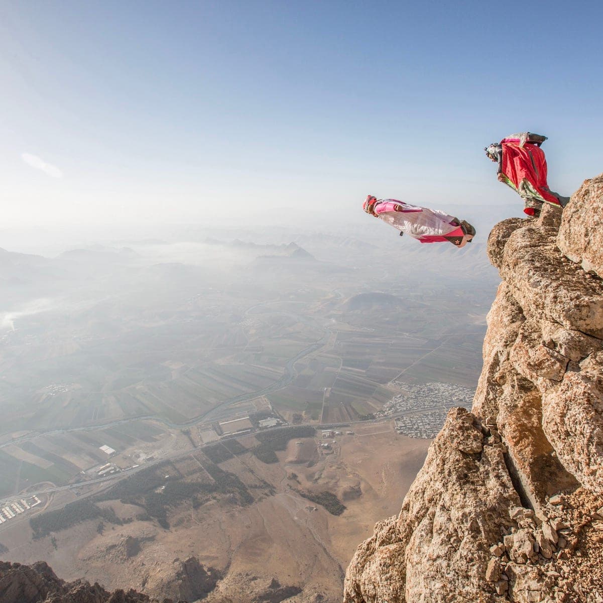 A wingsuiter in flight above a hazy valley, another person on the edge of a rocky spur, Bisotoun