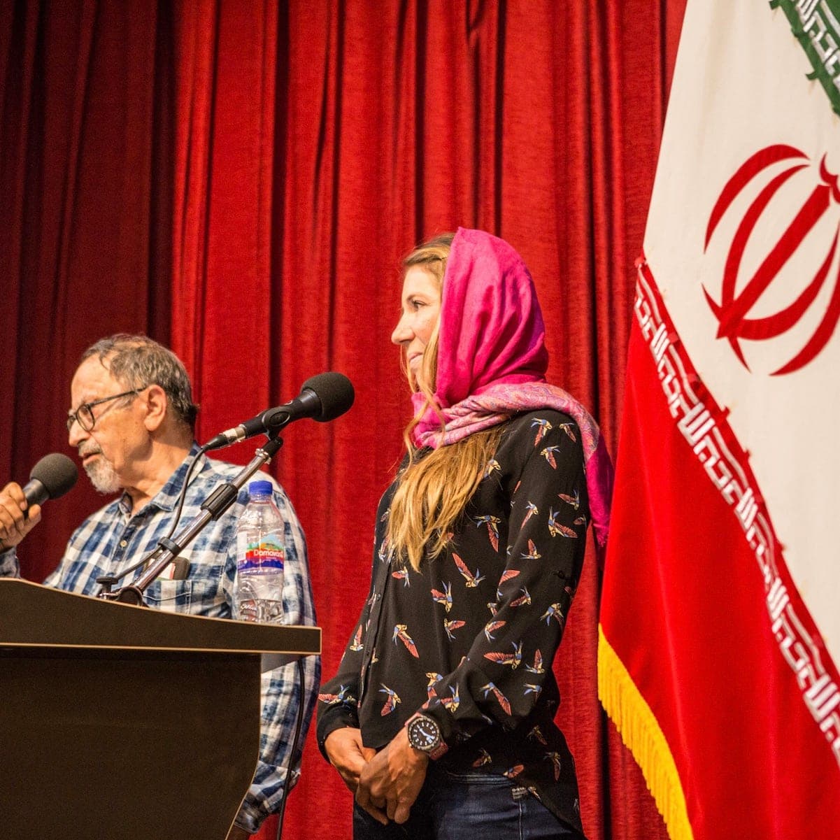 A man at a lectern and a woman in a pink headscarf on stage, beside the Iranian flag