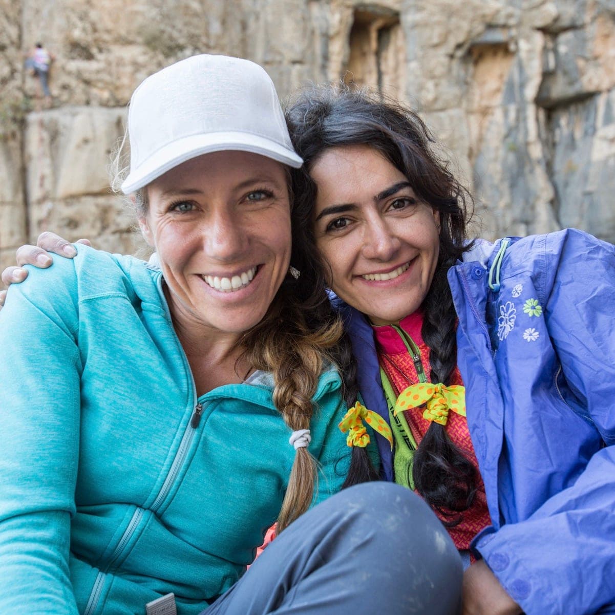 Two smiling women, one in a white cap and teal fleece, in front of a rock wall, Iran