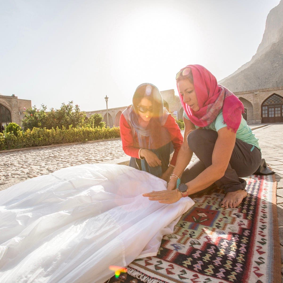 Two women in headscarves folding a large white canopy on a rug in a courtyard, Iran