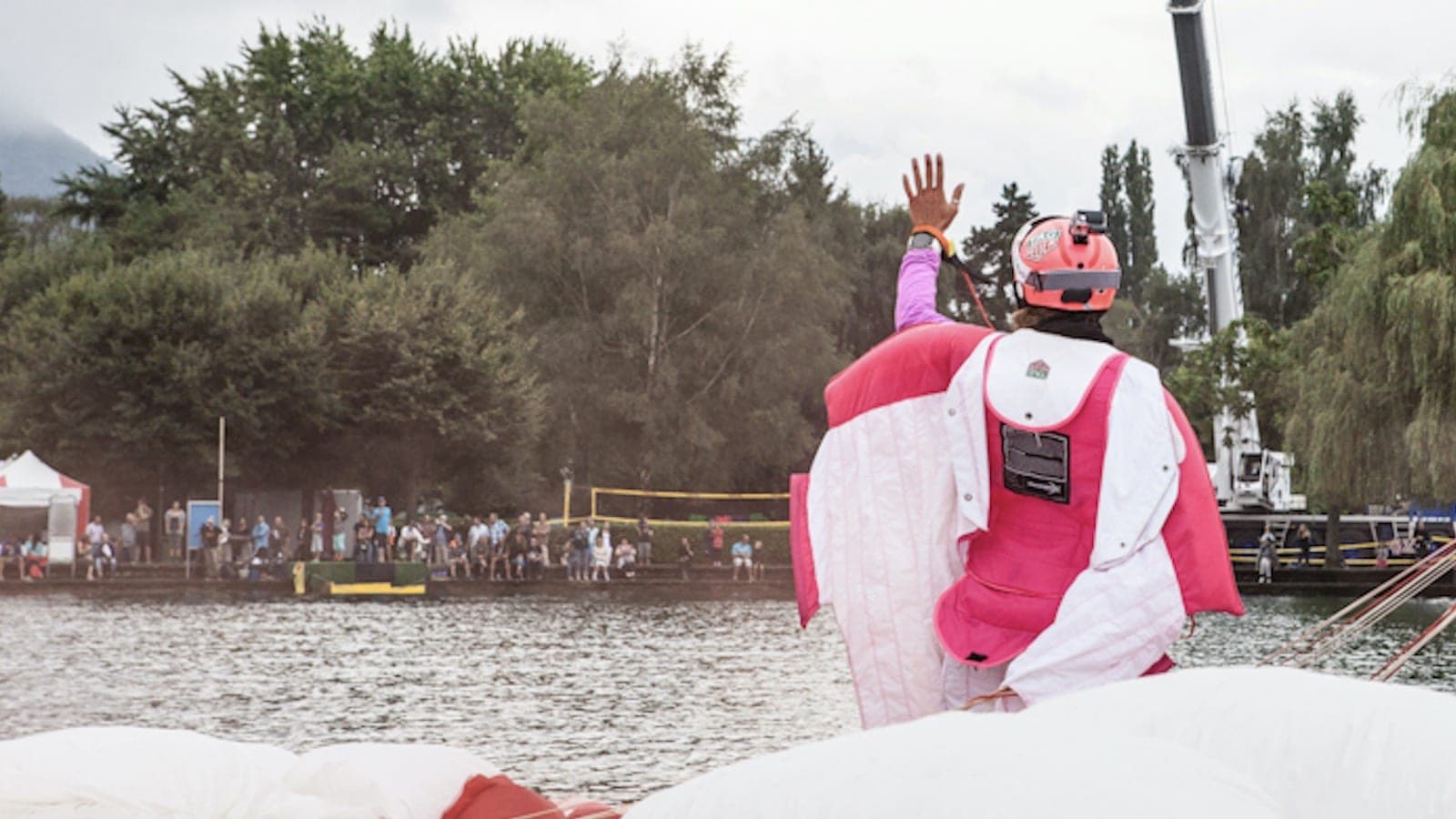 Un pilote de wingsuit rose et blanc, de dos, le bras levé, au bord d'un lac avec des spectateurs sur l'autre rive