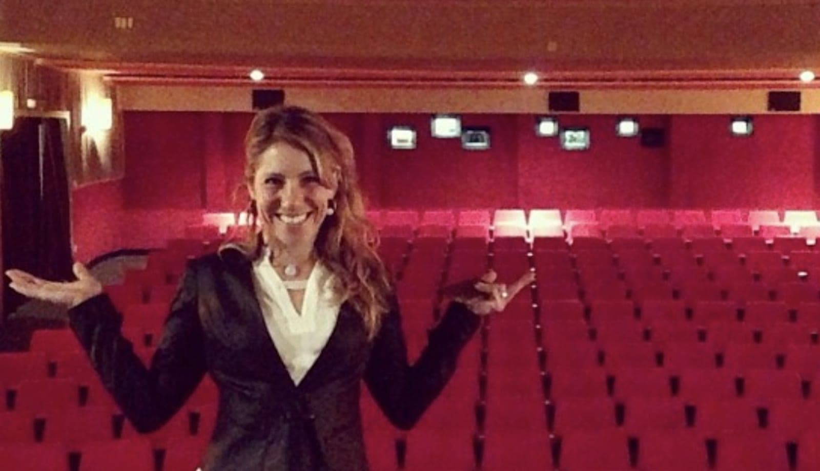 Une conférencière souriante, bras ouverts, devant les rangées de fauteuils rouges d'une salle de spectacle vide