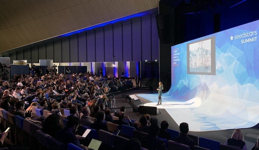 Une conférencière sur la scène du Seedstars Summit devant un large public, écran bleu à droite