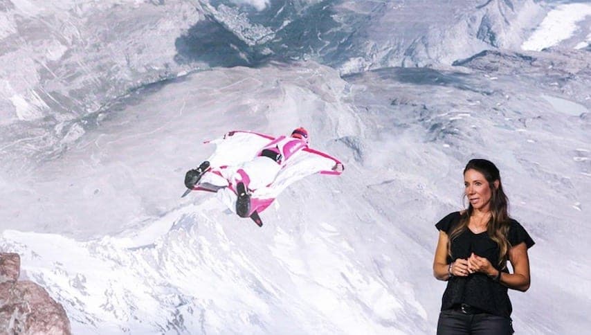 Une conférencière devant un écran montrant un pilote de wingsuit en vol au-dessus de montagnes