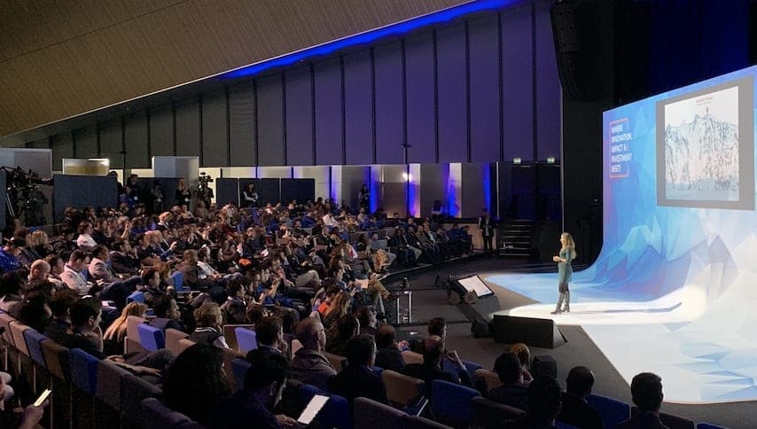 Une conférencière sur scène devant un large public dans une grande salle, écran bleu à droite