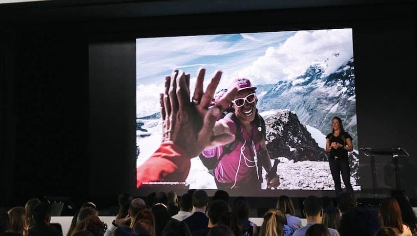 Une conférencière devant un écran où deux personnes se tapent dans la main au sommet d'une montagne, public au premier plan