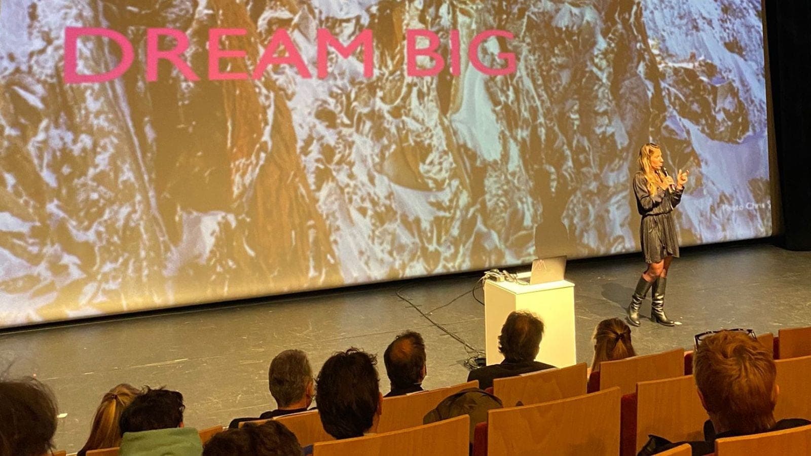 Une conférencière sur une scène devant un grand écran portant « Dream Big », public au premier plan