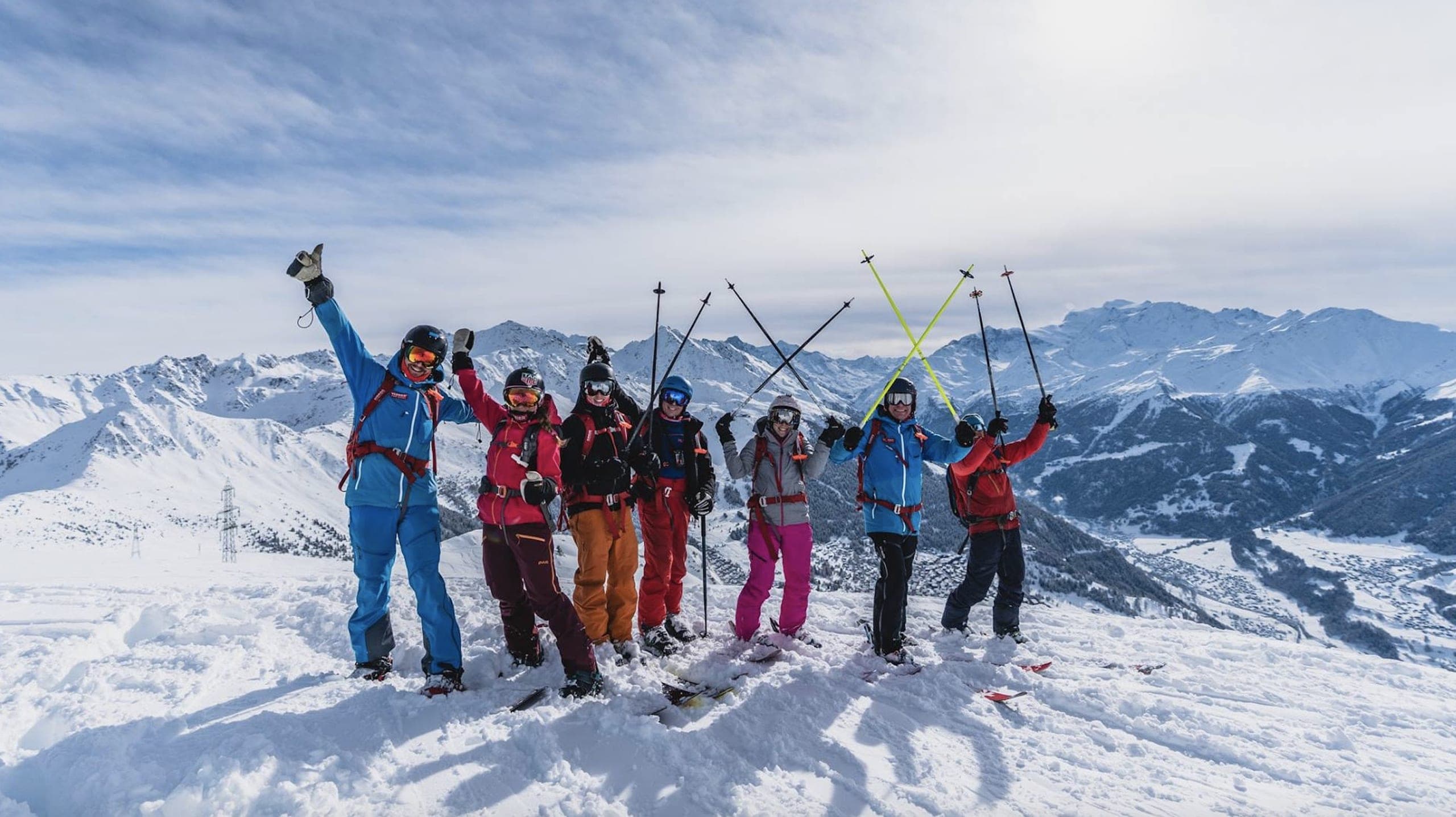 Un groupe de skieurs lève les bras et les bâtons au sommet d'une crête enneigée, montagnes derrière