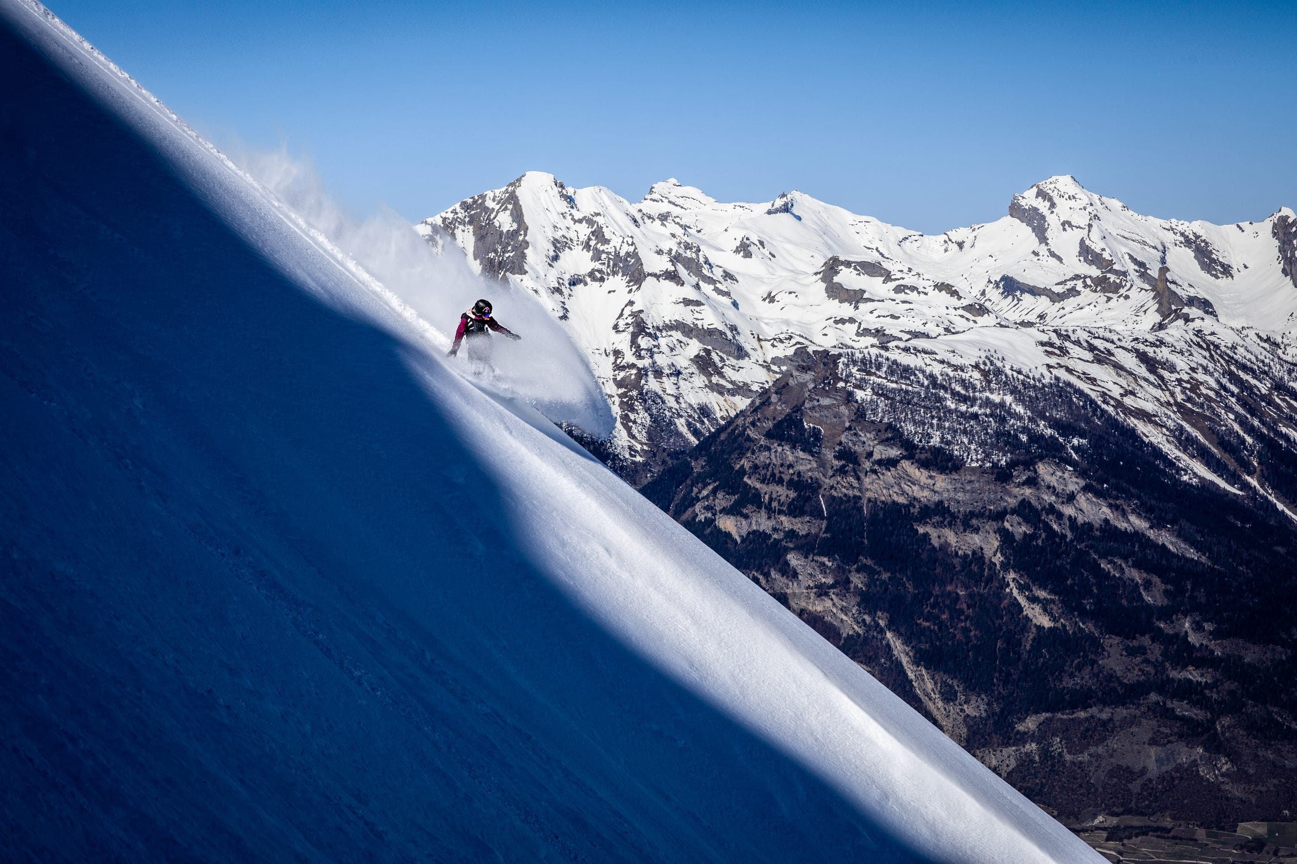 Une snowboardeuse descend une pente raide de poudreuse, face à des sommets enneigés