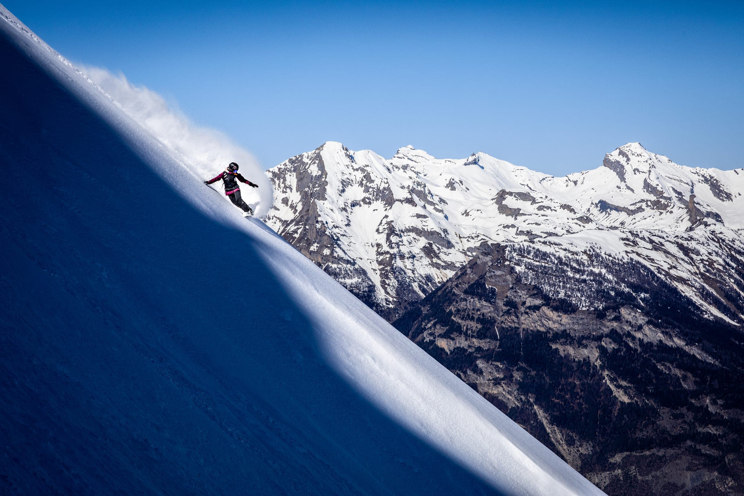 Une snowboardeuse soulève un nuage de neige sur une grande pente ombragée, devant des montagnes