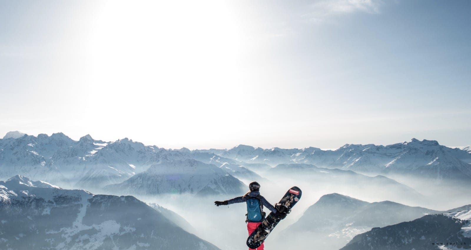 Une snowboardeuse de dos, planche sous le bras, face à des montagnes bleutées dans la brume