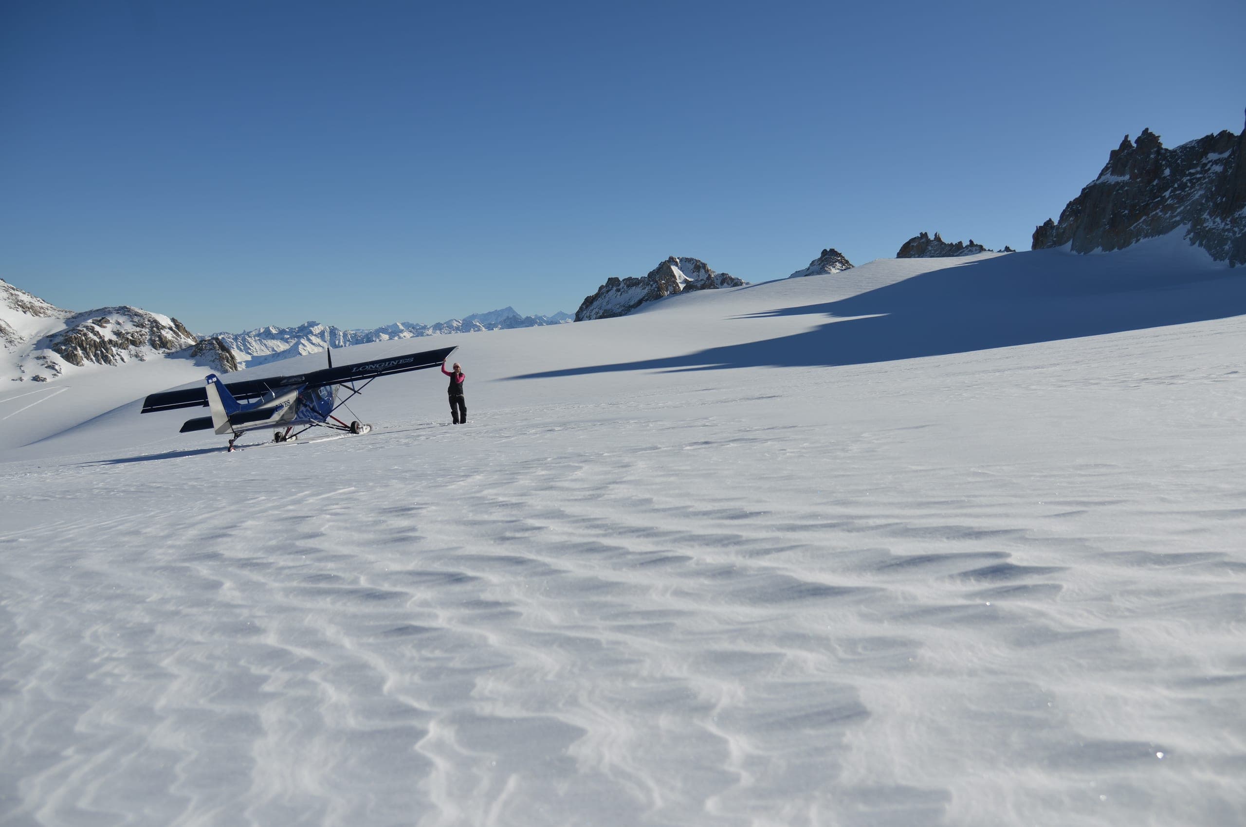 Un petit avion posé sur un glacier ondulé de neige, un pilote debout près de l'appareil