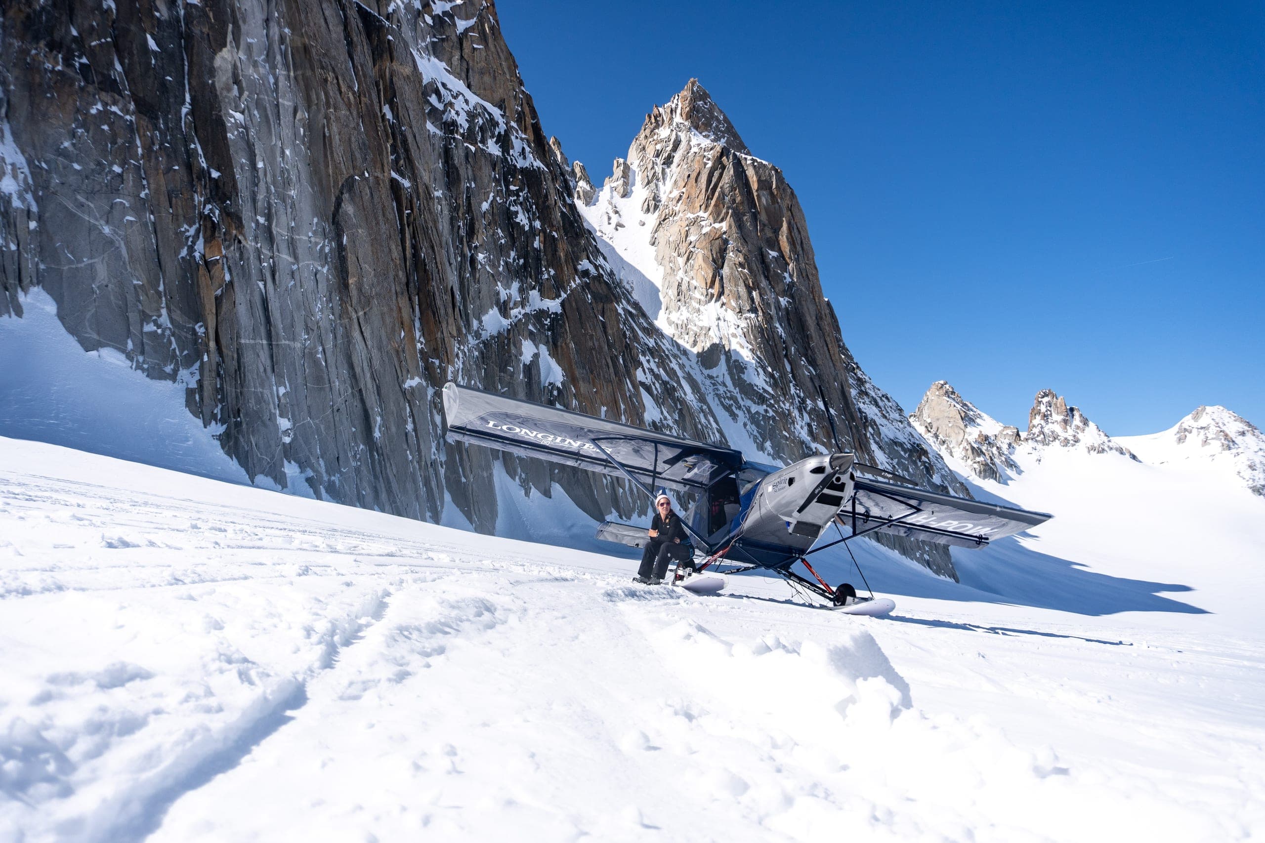 Un avion à skis posé sur un glacier au pied d'une paroi rocheuse et d'aiguilles, un pilote à côté