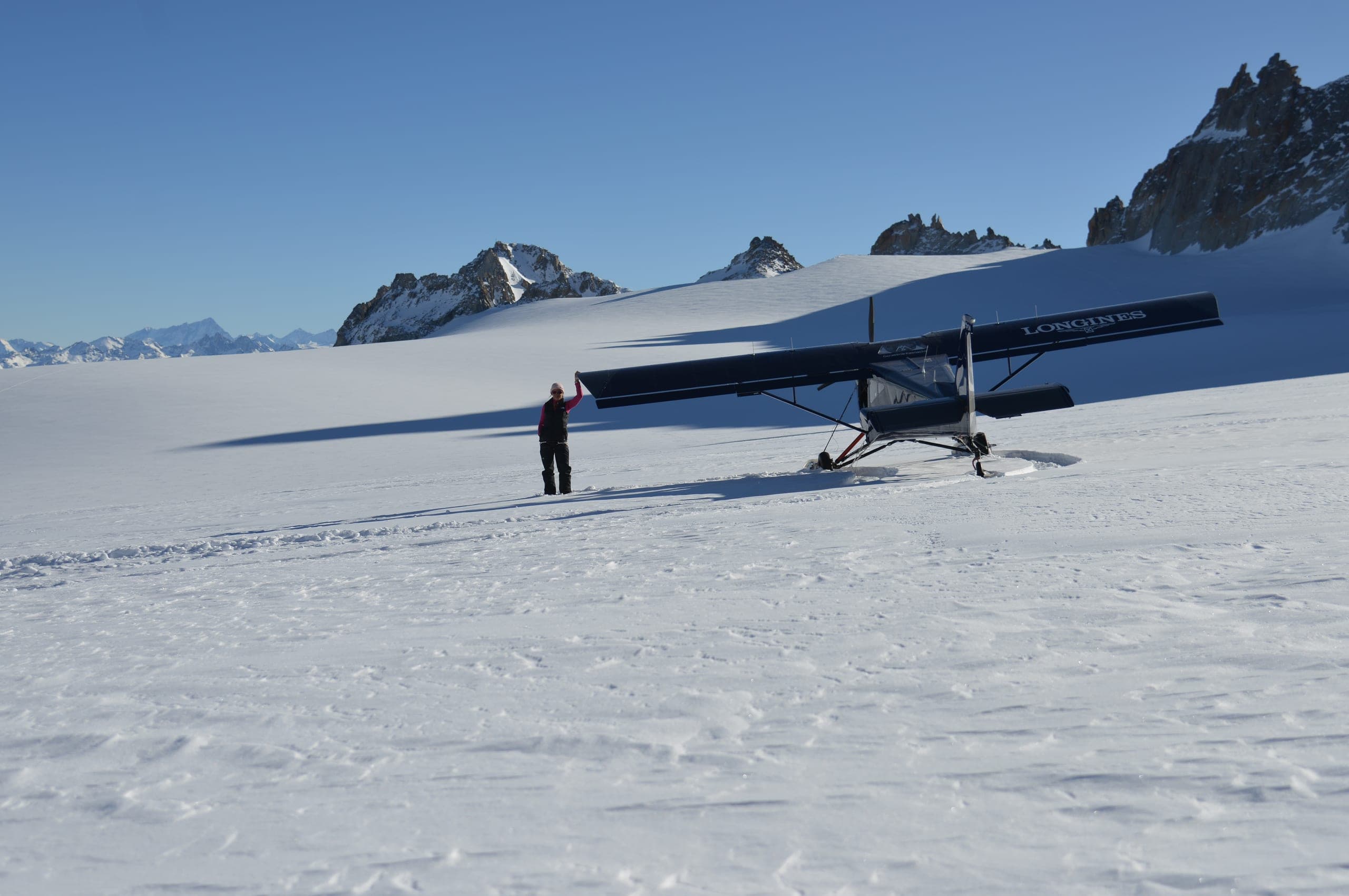 Un avion à skis posé sur un vaste glacier enneigé, un pilote debout à côté, sommets au loin