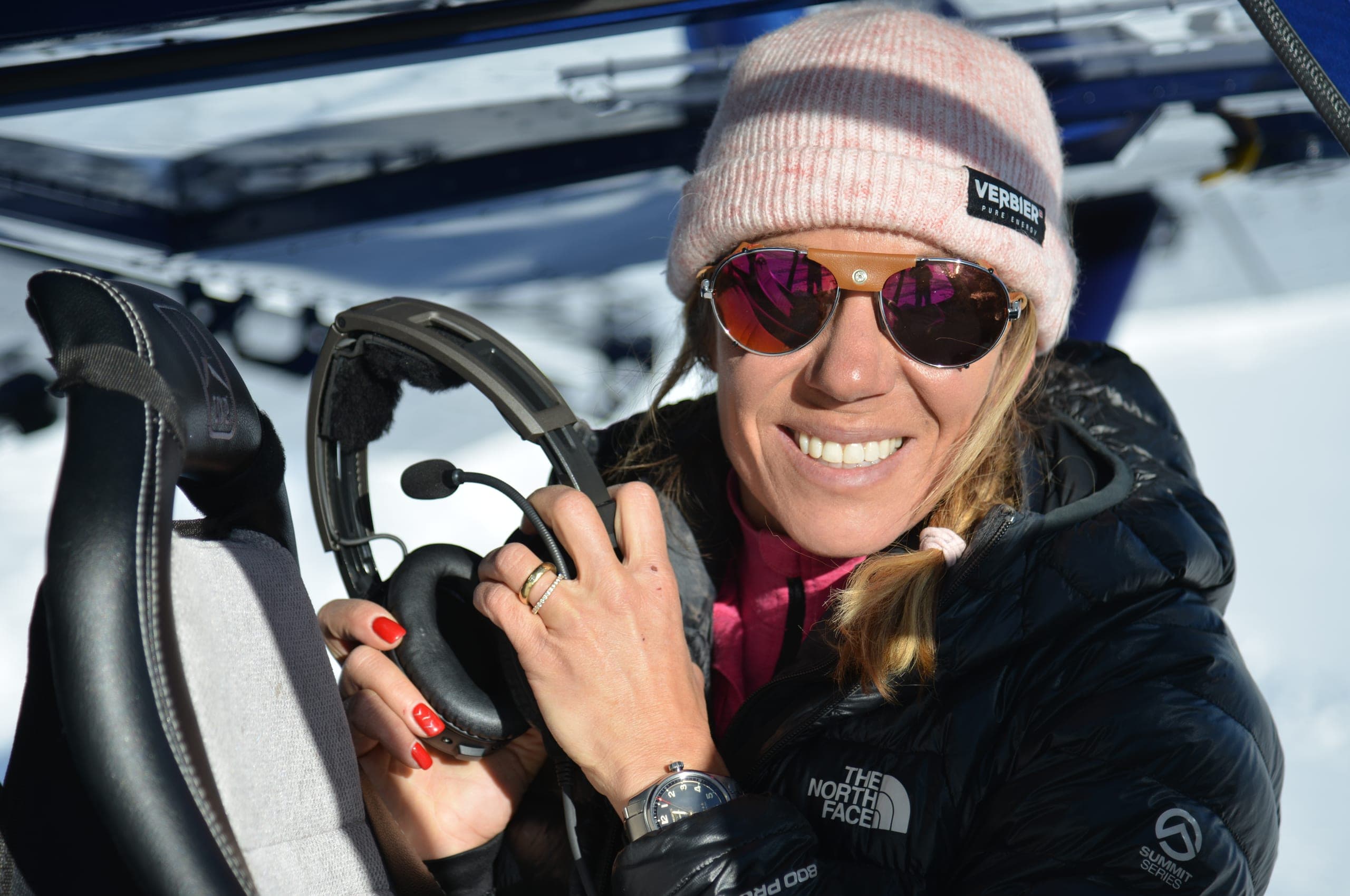 Une pilote souriante en bonnet rose, lunettes de soleil, tient un casque audio dans un cockpit