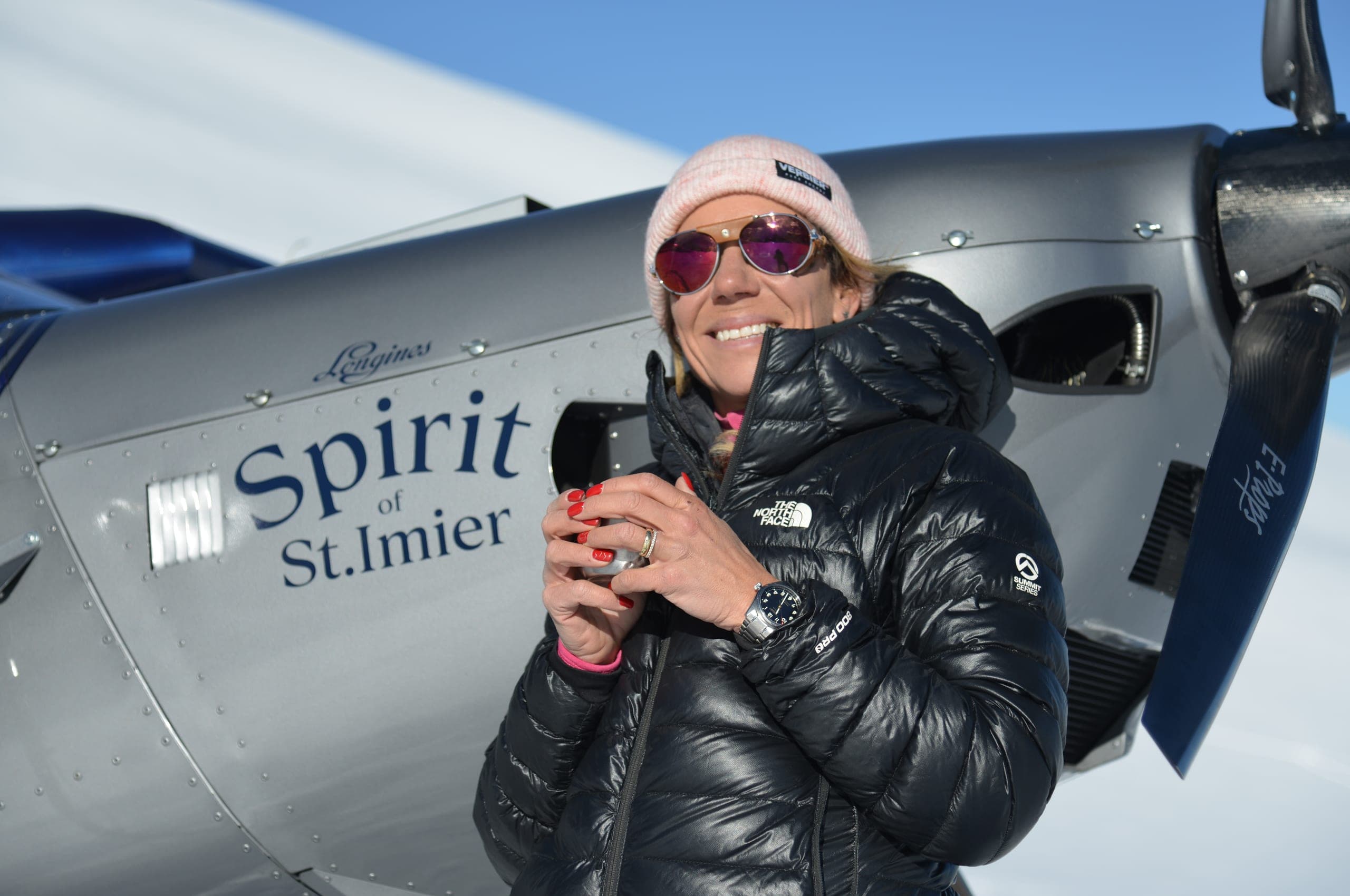Une pilote souriante devant l'hélice et le capot d'un avion gris marqué « Spirit of St.Imier »