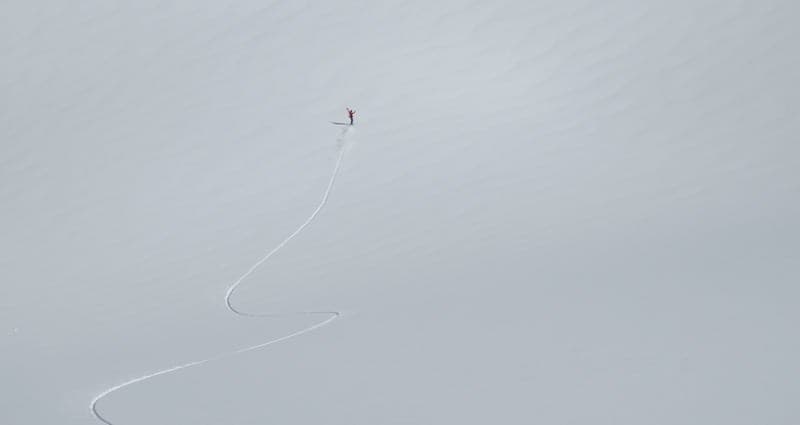Vue aérienne d'une grande étendue de neige où une trace sinueuse descend vers une minuscule silhouette