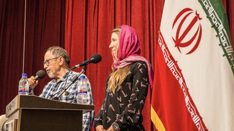 Un homme au micro et une femme en foulard rose sur une estrade, devant un rideau rouge et un drapeau iranien
