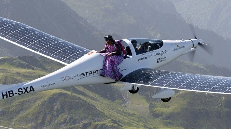 Une personne en tenue colorée assise sur l'aile d'un avion solaire immatriculé HB-SXA, au-dessus de collines vertes