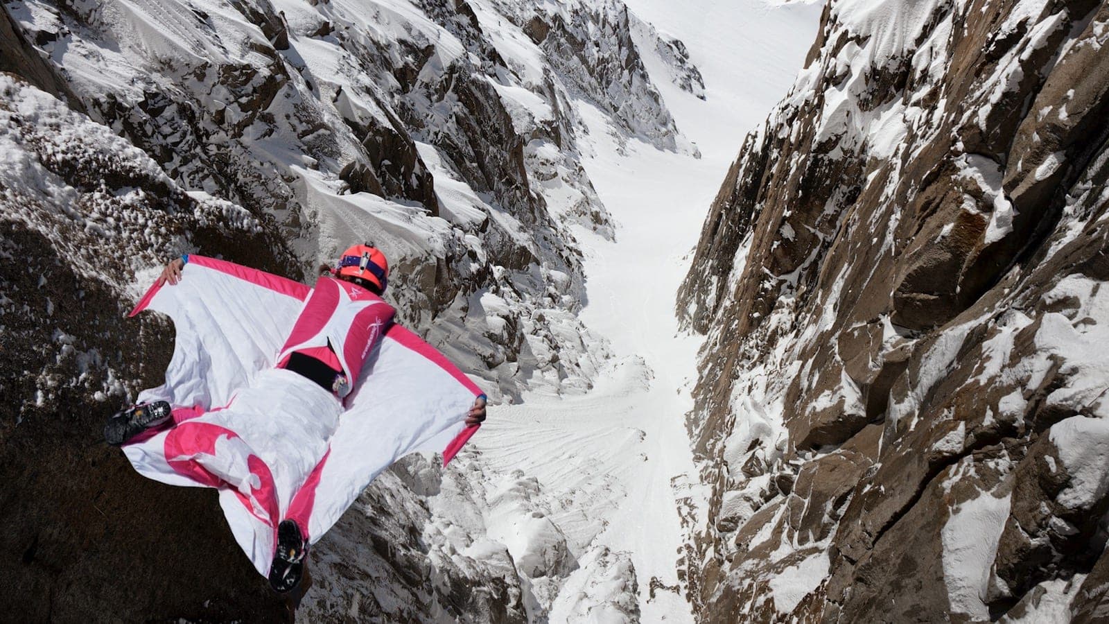 Un pilote en combinaison wingsuit blanche et rose vole au-dessus d'un couloir enneigé entre deux parois