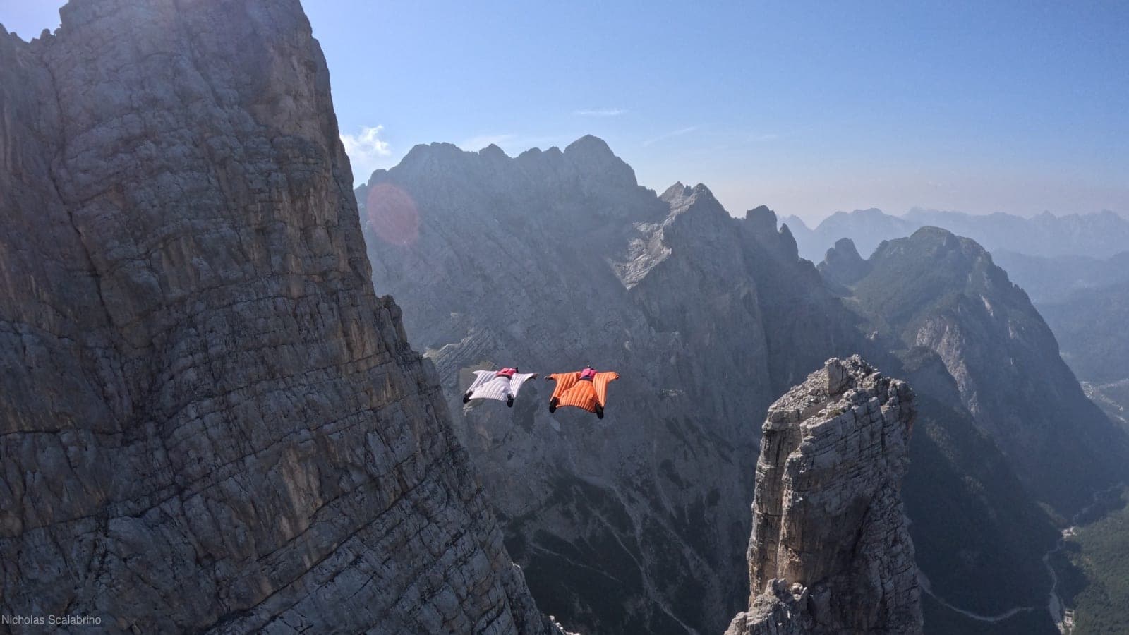 Deux pilotes de wingsuit, l'un en blanc et l'autre en orange, volent entre des parois et aiguilles rocheuses