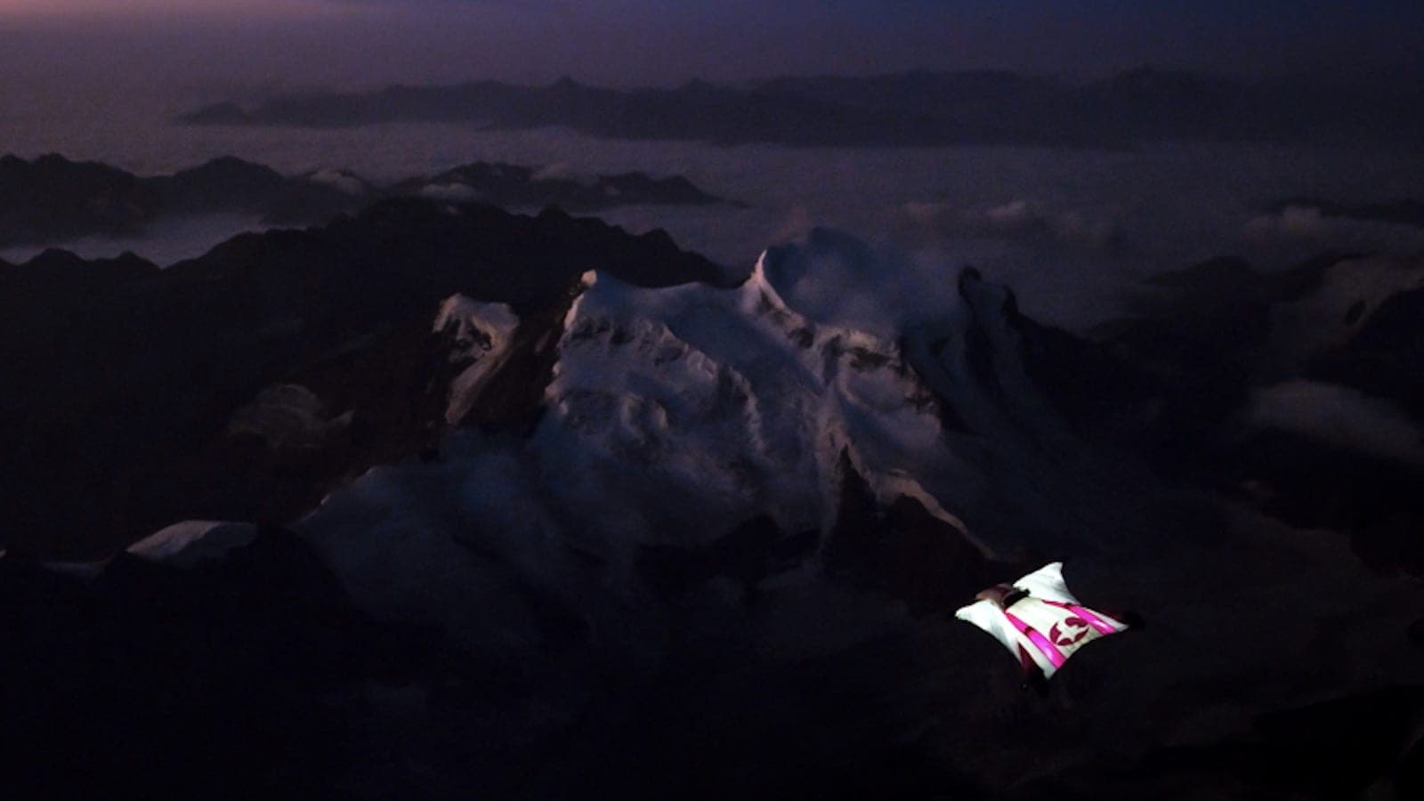 Un pilote de wingsuit en vol au crépuscule au-dessus d'un massif enneigé plongé dans l'ombre