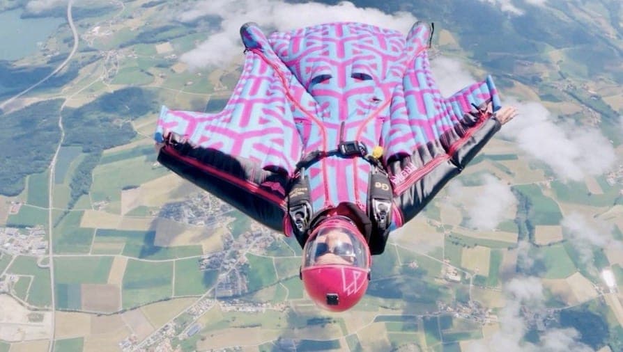 Une pilote de wingsuit à motifs bleus et roses vole au-dessus de champs verts et de nuages